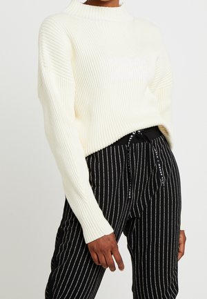 Personne portant un pull côtelé crème à manches longues et un pantalon noir à fines rayures verticales blanches avec une taille à cordon.