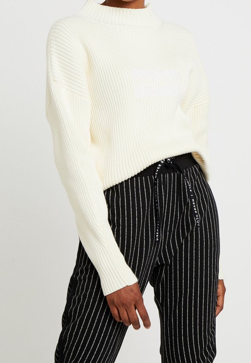 Personne portant un pull côtelé crème à manches longues et un pantalon noir à fines rayures verticales blanches avec une taille à cordon.