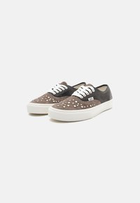 Brune semskede og sorte lærret sneakers med hvite snører, dekorativt hvitt prikkemønster, gummisåle, og Vans-logo på siden.