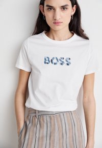 Balta medvilnės marškinėliai su tekstūruotu mėlynu "BOSS" užrašu. Modelis dėvi dryžuotas, aukštu juosmeniu kelnes su kišenėmis. Paprastas apvalus iškirpimas.