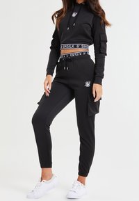 Sudadera recortada negra con un logo blanco, combinada con pantalones deportivos negros con bolsillos laterales, terminada con zapatillas blancas. Textura de tela estándar.