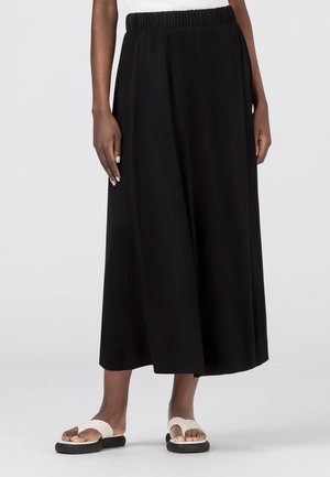 Personne portant un pantalon large noir court avec une taille élastique et des sandales beige avec des semelles noires sur fond gris.
