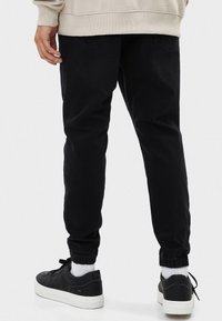 Pantalones de mezclilla negros con un ajuste tapered, puños elásticos y dos bolsillos traseros, llevados con zapatillas negras y calcetas blancas por los tobillos.