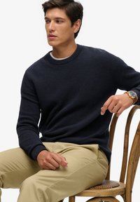 Marineblauwe textuursweater met een klassieke ronde hals, gecombineerd met lichtbeige broek. Model zit op een houten stoel.