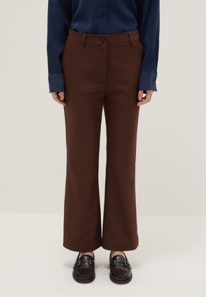 Personne portant un pantalon marron à jambes droites associé à des mocassins marron foncé et une chemise bleu marine à manches longues, debout devant un fond uni.