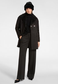 Donna con cappello di pelliccia nera, cappotto in shearling nero con colletto strutturato, dolcevita nera, pantaloni a righe con gamba larga e tacchi neri.