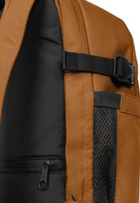 Hnědý látkový batoh s černým texturovaným panelem, síťovou kapsou a zipovanou přihrádkou; obsahuje robustní kovové komponenty a nastavitelné popruhy.