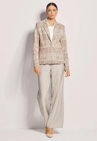 Licht geruite blazer in beige en bruin, met een enkele knoopsluiting, gecombineerd met lichtgrijze wijde broek en lichtbeige hakken.