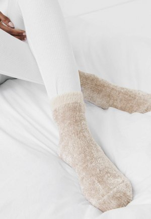 Chaussettes en beige flou avec une texture douce et un ourlet duveteux, assorties à des leggings blancs côtelés, reposant sur une surface blanche.
