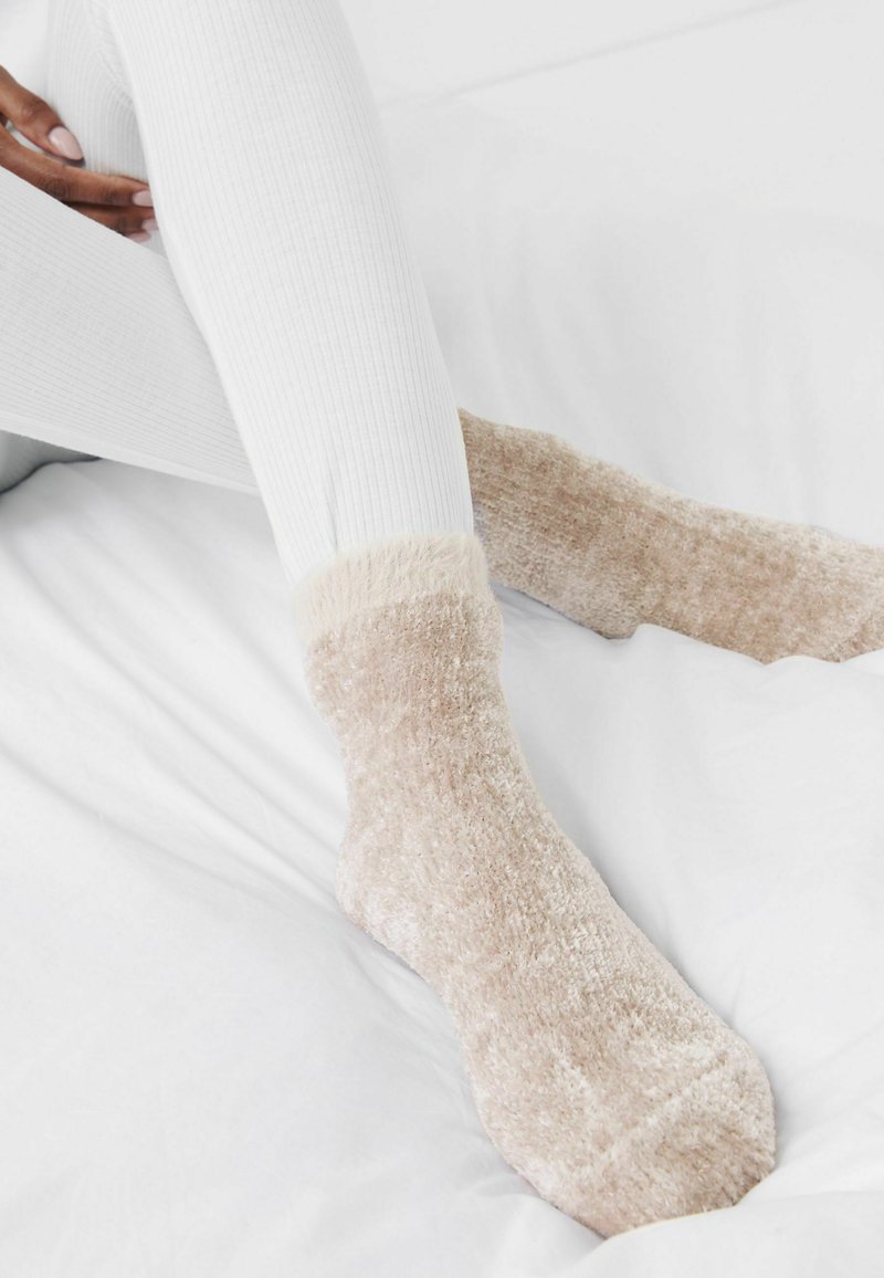 Flauschige beigefarbene Socken mit einer weichen Textur und einem flauschigen Bündchen, kombiniert mit weißen gerippten Leggings, die auf einer weißen Oberfläche liegen.