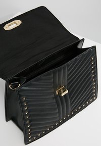 Svart faux läderhandväska med kviltad textur, gyllene metallaccenter och pyramider studs längs kanterna. Öppen klaffdesign.