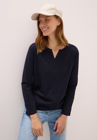 Marineblaues Langarmshirt mit V-Ausschnitt und subtilen Nähten, kombiniert mit hellblauen Jeans und einer hellen Kappe.