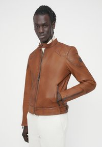 Καφέ δερμάτινο μπουφάν με εφαρμοστή γραμμή, κεντημένα διακοσμητικά στους ώμους, ψηλό γιακά και τσέπες με φερμουάρ. Λείο υλικό, μινιμαλιστικό στυλ.