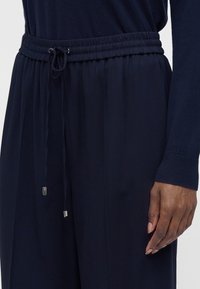 Personne portant un pantalon à taille élastique bleu marine foncé avec un cordon de serrage et un haut à manches longues assorti, main détendue le long du corps.