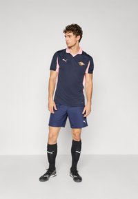 Tamsiai mėlyna futbolo apranga su rožinėmis detalėmis, trumpomis rankovėmis ir apykakle. Derinama su tamsiai mėlynais šortais ir juodomis kojinėmis. Aprangoje matomas Puma logotipas.