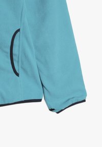 Fleecejakke i lys blå med en glat tekstur. Har sorte kanter og en sidelomme. Ærmerne er kantede, og designet er afslappet.