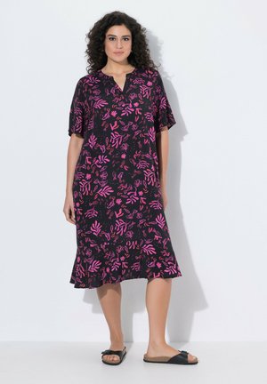 Vrouw met krullend donker haar die een zwarte midijurk met roze botanische patronen en zwarte slidesandalen draagt, staand tegen een witte achtergrond.