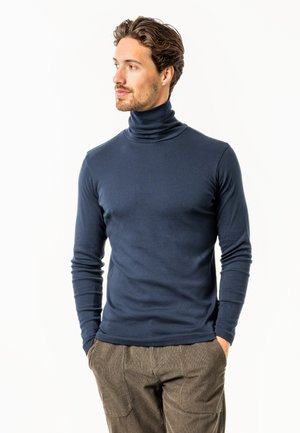 Mann in einem marineblauen Rollkragenpullover und brauner Hose, steht mit den Händen in den Taschen und blickt nach links.
