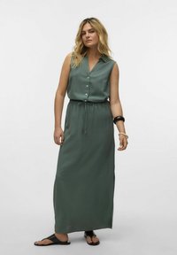 Robe maxi sans manches, vert olive, avec un devant à boutons, une taille ajustable par cordon et des fentes latérales. Portée avec des sandales noires.