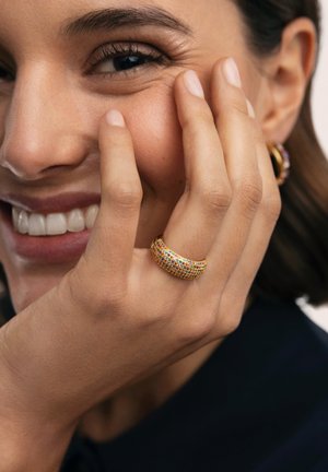 Anillo de oro con piedras preciosas multicolores, con un diseño liso y curvado. El modelo se toca la cara, mostrando pendientes ornamentados en el fondo.