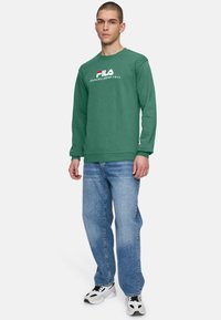 Groene sweatshirt met lange mouwen en het witte FILA-logo, gecombineerd met lichtblauwe rechte spijkerbroeken en zwart-witte sneakers.