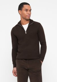 Ruda zip-up megztinis su tekstūriniu mezgimu, turintis aukštą apykaklę ir sidabrinę užtrauktuką. Suderintas su rudais tekstūriniais kelnėmis.