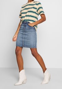 Randig beige och grön bomullströja nedstoppad i en knälång blå denimkjol, bärs med vita klackiga cowboystövlar.