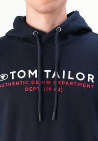 Granatowy hoodie z sznurkiem i srebrnymi metalowymi końcówkami. Z przodu znajduje się biały i czerwony tekst: "TOM TAILOR AUTHENTIC DENIM DEPARTMENT."