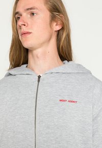 Szara bluza z kapturem na zamek, o miękkiej teksturze. Czerwony haft "NIGHT ADDICT" na lewej piersi. Regulowany kaptur.