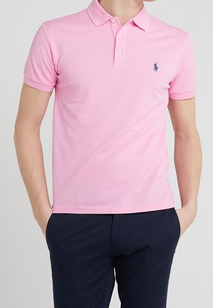 Homme portant un polo rose clair à manches courtes avec un petit logo foncé sur la poitrine et un pantalon foncé, les mains dans les poches.