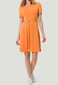 Pomarańczowa sukienka z krótkim rękawem, z paskiem w talii, długości do kolan, z delikatnymi rozcięciami po bokach. W zestawie z białymi sneakersami dla uzyskania casualowego wyglądu.