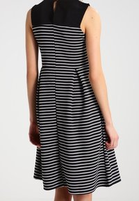 Kvinna i en ärmlös svart klänning med vita horisontella ränder och en enfärgad svart övre del, stående mot en enkel bakgrund.