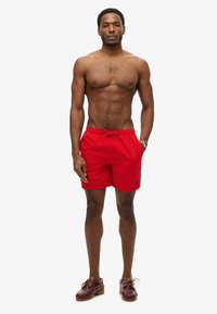 Rode zwemshorts met een elastische tailleband en een trekkoord. Het model draagt bruine bootschoenen, die een eenvoudige design en een knie-lange snit tonen.