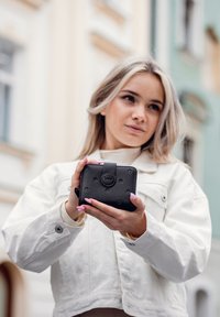 Černá peněženka s kruhovými vzory a zapínáním na cvok, držená v rukou. Na přední straně je emblém značky. Pozadí tvoří světlá architektura.