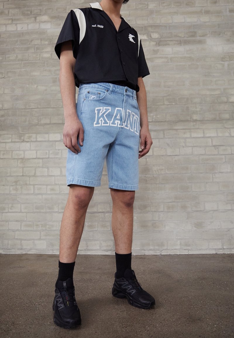 Niebieskie dżinsowe szorty z białym napisem "KANI", wysoki stan i lekko postrzępione wykończenie. Połączone z czarną koszulą z krótkim rękawem i czarnymi sneakersami.