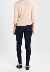 Beige gebreide trui met driekwart mouwen, gecombineerd met nauwsluitende donkerblauwe jeans. De outfit omvat simpele zwarte ballerina's.