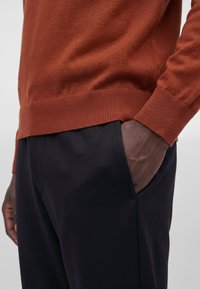 Personne portant un pull couleur rouille avec une main dans la poche du pantalon noir, montré de la taille à mi-cuisse.