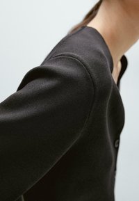 Cardigan nero lavorato a maglia con scollatura rotonda, chiusura a bottoni e superficie testurizzata con spalle leggermente imbottite.