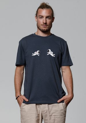 Navy blue cotton t-shirt featuring two white astronaut graphics, a starry background, and short sleeves. Main design centered on chest.