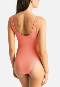 Maillot de bain une pièce en tissu côtelé de couleur corail, avec des bretelles larges et un dos bas. La coupe est ajustée avec un design de jambe haute.