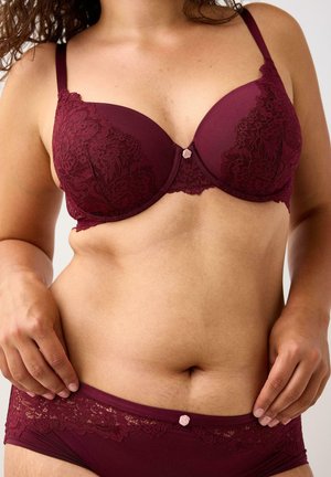 Donna che indossa un reggiseno e slip in pizzo borgogna abbinati, mentre sistema il pantaloncino, su uno sfondo neutro.