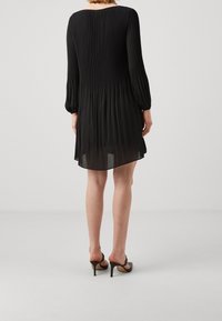 Robe noire plissée avec des manches longues et un dos fluide. Tissu léger et transparent. Associée à des talons hauts ouverts.