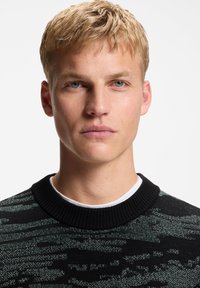 Νέος άνδρας με κοντά ξανθά μαλλιά φοράει μαύρο και πράσινο πουλόβερ με μοτίβο πάνω από λευκό πουκάμισο, κοιτάζοντας κατευθείαν την κάμερα σε λευκό φόντο.