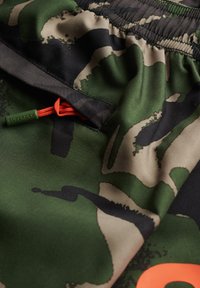 Shorts en camouflage vert avec des accents noirs, dotés d'un cordon de serrage orange et d'une étiquette portant la mention "Superdry". Tissu lisse et léger.