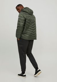 Chaqueta acolchada de color verde con capucha, con un patrón en forma de Chevron. Se lleva con pantalones negros ajustados y zapatillas negras.