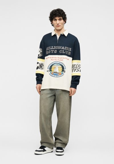 Νέος άντρας που φοράει μπλούζα rugby μακρυμάνικη Billionaire Boys Club με ναυτικά λογότυπα, χαλαρό γκρι παντελόνι και μαύρα-άσπρα αθλητικά παπούτσια.