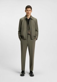 Olijfgroene jas en broek van een gladde stof. De jas heeft een kraag en een elastische tailleband, gecombineerd met een zwart shirt eronder.