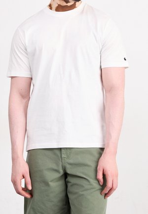 Man die een effen wit T-shirt met korte mouwen en olijfgroene broek draagt, staand tegen een witte achtergrond.