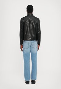 Melna ādas jakas ar spiedpogu apkakli, pieguļošs dizains un redzamas šuves, kombinēta ar gaiši ziliem taisnajiem džinsiem un melnām kurpēm.