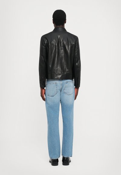 Sort læderjakke med knapkrave, tætsiddende design og synlige sting, parret med lyseblå lige ben jeans og sorte sko.